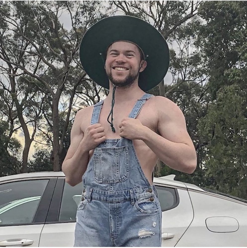 Me, shirtless under denim overalls and a wide-brimmed hat, grinning in front of a white car under gum trees